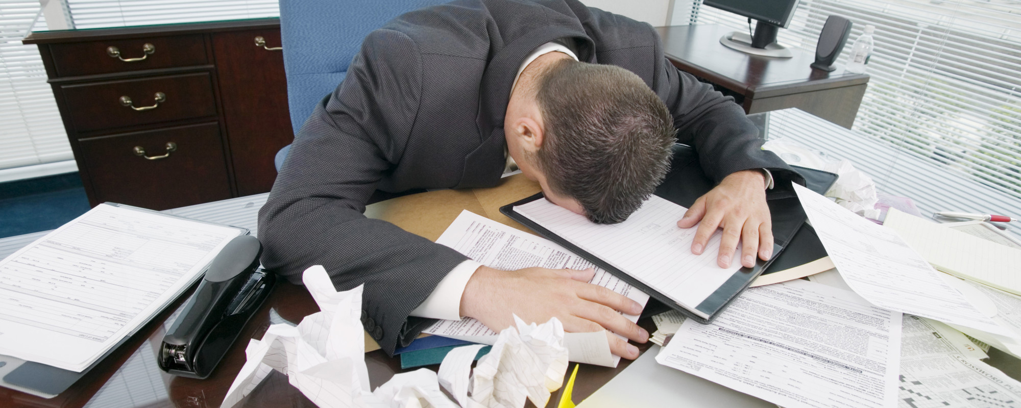 A man in a suit leans forward with his head down on a cluttered desk, covered with papers and crumpled documents, reflecting the overwhelming demands of people management in an office setting. A man in a suit leans forward with his head down on a cluttered desk, covered with papers and crumpled documents, reflecting the overwhelming demands of people management in an office setting.